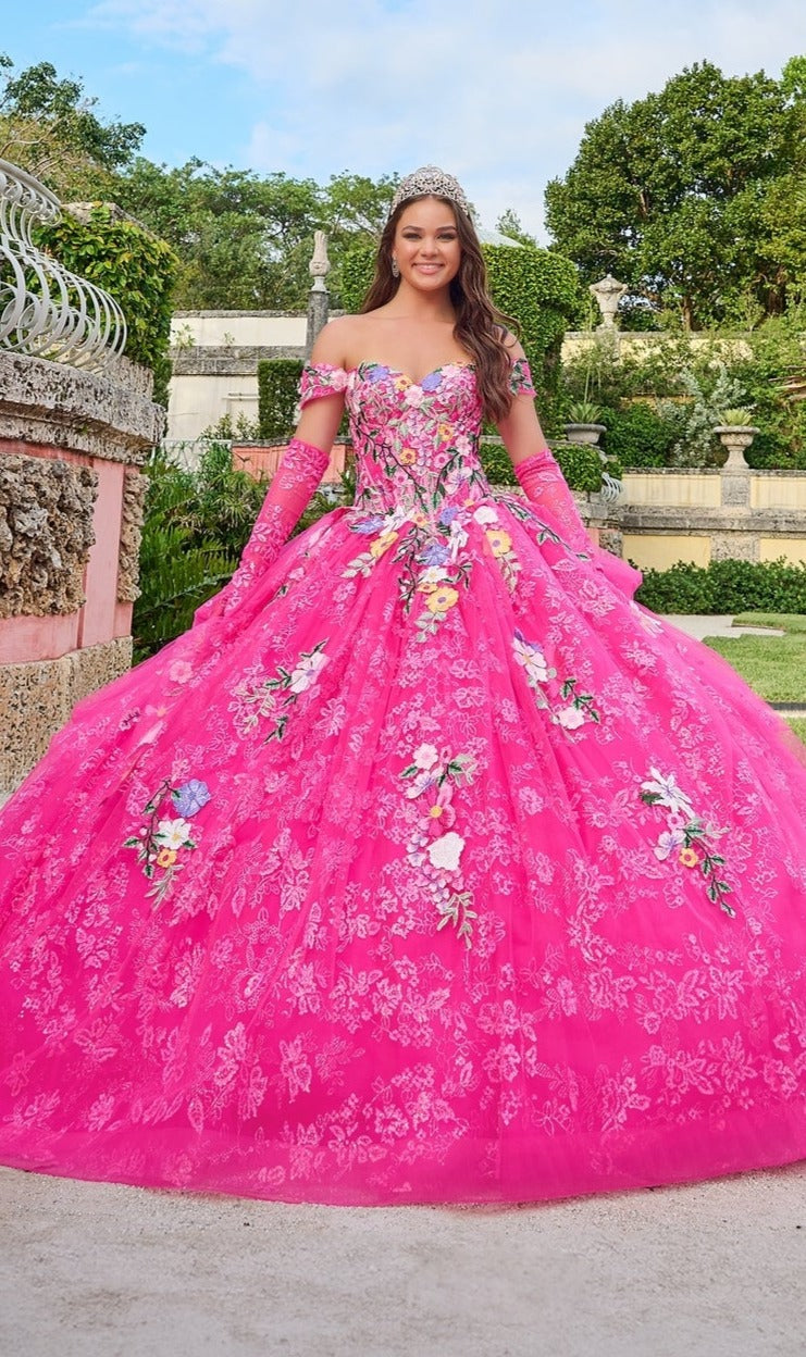 A young woman wearing the Quinceanera Dress 54200 By Amarra, a bright pink off-the-shoulder gown with floral embroidery and a tiara, stands smiling in a garden.